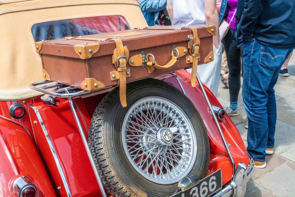 A leather suitcase strapped above the rear-mounted spare wheel of a vintage red sports car
