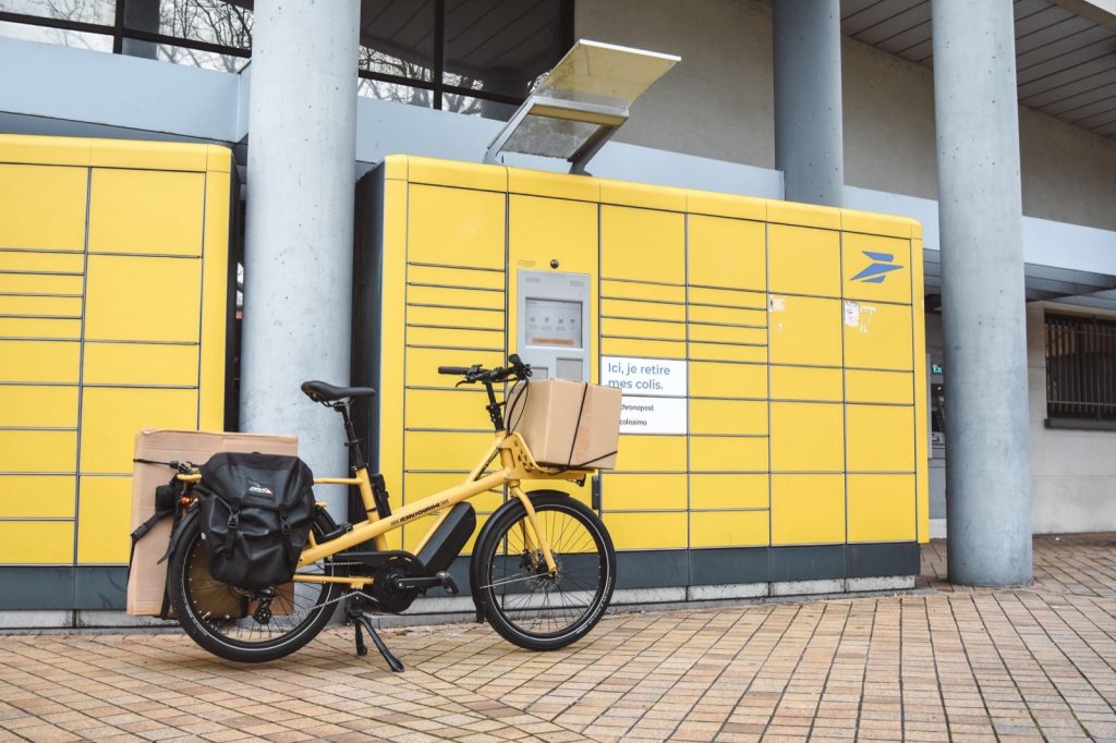 A yellow cargo bicycle loaded with parcels parked in front of a bank of yellow parcel lockers