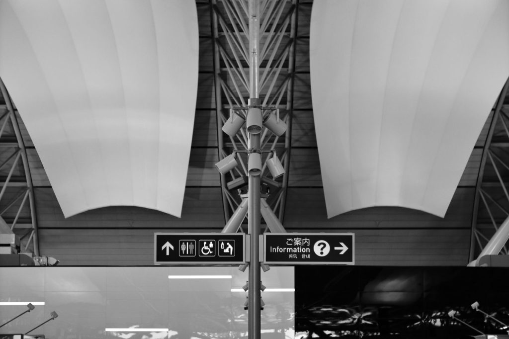 Overhead airport wayfinding signs with directional arrows and a multilingual information sign, in black and white
