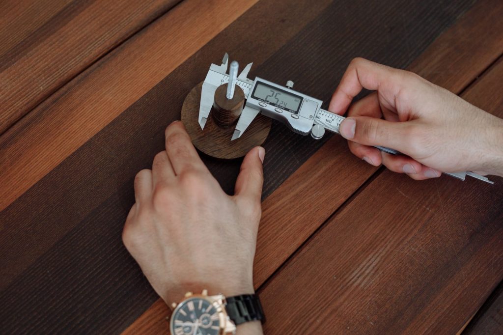Two hands using a digital vernier caliper to measure a small round wooden part on a dark wooden table