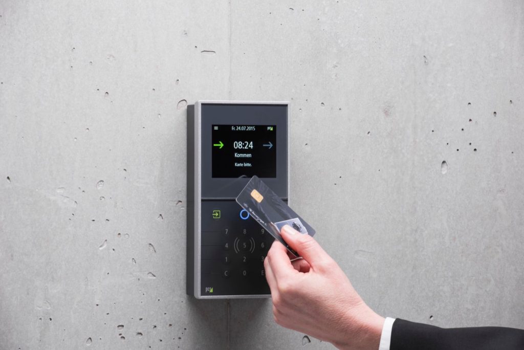 A hand holds an access card up to a wall-mounted card reader terminal on a concrete wall