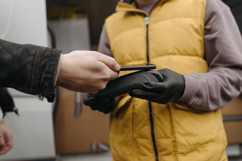 A customer taps a phone against a card terminal held by a courier in a yellow vest beside a delivery van