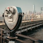 A coin-operated viewing machine on a wooden pier aimed at a distant city skyline softened by haze