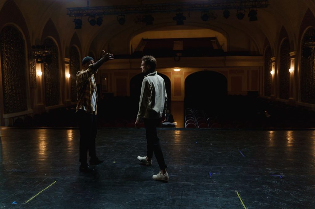 Two people rehearsing on a dimly lit theatre stage with marking tape on the floor and an empty auditorium behind